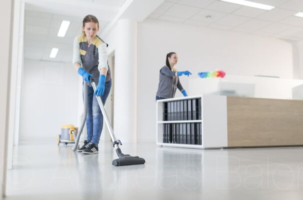 Eine Mitarbeiterin saugt den Boden in einem Büro während eine andere Dame im Hintergrund Staub wischt.