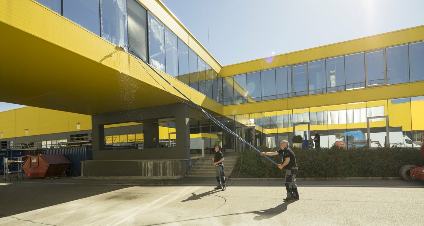 Zwei Arbeiter putzen ein Fenster einer Glasfront über ihnen mit einer ausfahrbaren Bürste