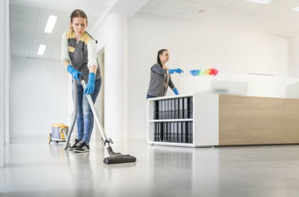 Eine Mitarbeiterin saugt den Boden in einem Büro während eine andere Dame im Hintergrund Staub wischt.